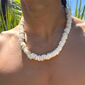May include: A white shell necklace composed of numerous small, flat, circular shells strung together. The necklace is worn around the neck, with a natural outdoor background of green foliage and blue sky.