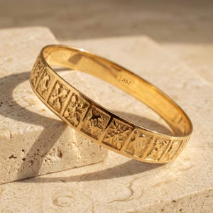 May include: A gold-colored bangle bracelet with a series of embossed square designs. The bracelet has a smooth, polished finish and the text "14K" is visible. The bracelet is displayed on a light-colored, textured surface.