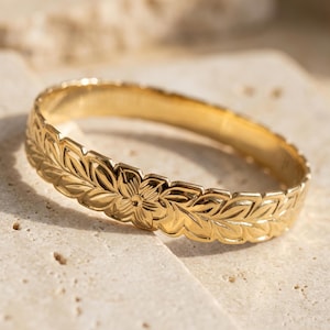 Op de afbeelding: Een goudkleurige armband met een bloemen- en bladerenpatroon. De armband heeft een gedetailleerd ontwerp van bloemen en bladeren die rond de hele band zijn gesneden. De sieraden worden gepresenteerd op een lichtgekleurd, getextureerd oppervlak.