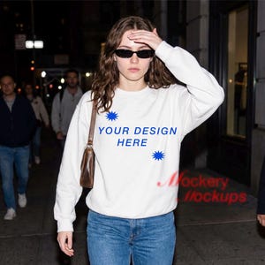 May include: White sweatshirt with the text "YOUR DESIGN HERE" in blue, along with blue starburst graphics. The sweatshirt is worn with blue jeans and sunglasses. The image is taken outdoors at night.