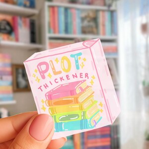 May include: A square sticker featuring a pastel pink border and the text PLOT THICKENER in colorful letters. The design includes a stack of five books in pink, orange, yellow, green, and blue, accented with small yellow sparkles on a white background.