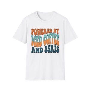 Peut inclure: T-shirt blanc avec l'inscription "POWERED BY ICED COFFEE AND SSRIS" en orange, bleu et marine. Le t-shirt a un col rond et des manches courtes. Le texte est dans une police rétro.