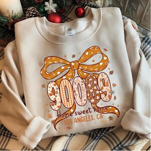 May include: A beige crewneck sweatshirt featuring a large orange bow graphic above the numbers 90089. The design includes autumn-themed patterns, leaves, and the text "Home sweet home LOS ANGELES, CA" printed in a dark brown script and block font.