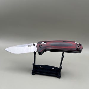 May include: A folding pocket knife with a silver-toned metal blade and a dark red and black wood-grain handle. The blade features a small butterfly logo and a thumb stud. The knife is positioned horizontally on a black plastic display stand.