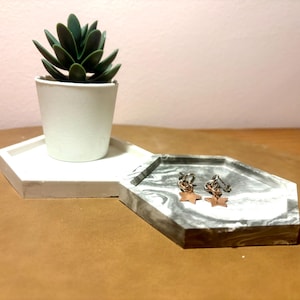May include: A white hexagonal tray holds a small white pot with a green succulent. A second tray, with a gray marble design, displays two pairs of star-shaped earrings. The items are on a brown surface.