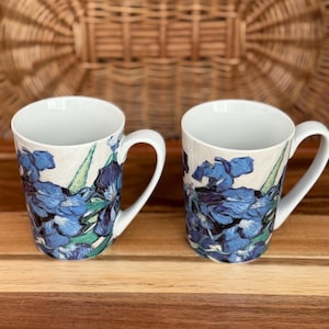 Può includere: Due tazze in ceramica bianca con un vivace motivo floreale di iris blu con foglie verdi. Ogni tazza ha un corpo cilindrico liscio, un manico curvo sul lato e un interno bianco semplice. Il design esterno mostra pennellate dettagliate.