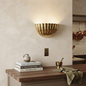 May include: A gold-toned, semi-circular wall sconce with a scalloped, shell-like design mounted on a wall. Below it, a stack of three books titled Salt, Fat, Acid, Heat, The Kitchen Muse, and Sourdough sits next to a small ceramic pitcher.