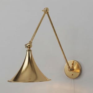Può includere: Una lampada da parete color oro con paralume conico e braccio articolato regolabile. L'apparecchio include una piastra a muro circolare e una finitura metallica riflettente, con un design snodato che consente un posizionamento direzionale.