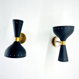 May include: Two wall sconces featuring matte black hourglass-shaped shades with small white perforated dots. Each lamp has a central gold-toned metal band and a circular gold-toned wall mounting plate, creating a symmetrical lighting fixture design.