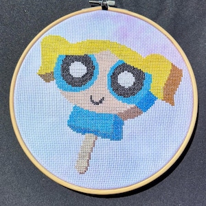 May include: Cross-stitch embroidery of a cartoon character in a wooden hoop. The character has blonde hair, large black eyes with blue accents, and a blue dress. The background is a light blue and purple watercolor effect.