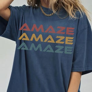 May include: A dark blue short-sleeved t-shirt featuring the word AMAZE printed three times in a stacked design. The top word is red, the middle is yellow, and the bottom is green. Each word has a distressed texture and a bold, sans-serif font.