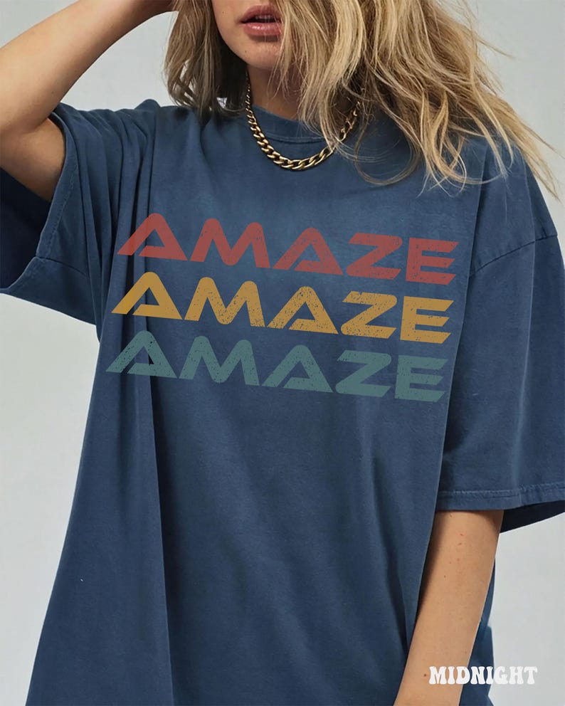 May include: A dark blue short-sleeved t-shirt featuring the word AMAZE printed three times in a stacked design. The top word is red, the middle is yellow, and the bottom is green. Each word has a distressed texture and a bold, sans-serif font.