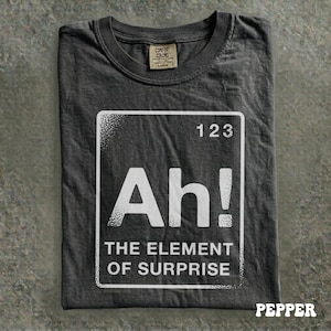 May include: Dark gray t-shirt with a white graphic that reads "Ah! THE ELEMENT OF SURPRISE" inside a square with the number "123" at the top. The shirt has a Comfort Colors tag.