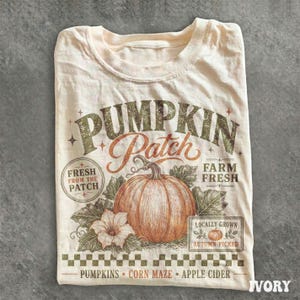 May include: An ivory t-shirt featuring a large orange pumpkin illustration with green leaves and a flower. Text reads PUMPKIN PATCH, FRESH FROM THE PATCH, FARM FRESH, LOCALLY GROWN, AUTUMN PICKED, PUMPKINS, CORN MAZE, and APPLE CIDER with a checkered border.