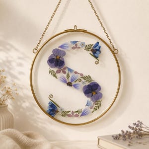 May include: A circular brass frame hanging by a gold chain, enclosing a clear glass pane. Pressed purple and blue pansies, small light blue flowers, and green fern leaves are arranged to form the letter S inside the circular glass display.