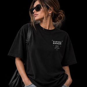 May include: A black short-sleeved t-shirt featuring white text on the upper left chest that reads SYSTEM ERROR 404 IDENTITY NOT FOUND, enclosed within thin white corner brackets and a horizontal line below the text.