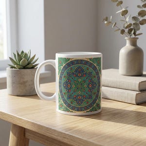 Op de afbeelding: Witte keramische mok met een gedetailleerd, cirkelvormig ontwerp in teal, blauw en rood. Het patroon doet denken aan traditioneel tegelwerk. De mok heeft een wit oor en staat op een houten oppervlak.
