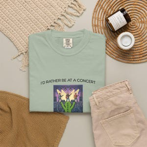 Peut inclure: Un t-shirt vert clair avec l'inscription "I'D RATHER BE AT A CONCERT" au-dessus d'un dessin d'un groupe jouant sur scène. Le t-shirt est présenté avec un pantalon beige et un tapis tissé.