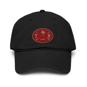 May include: A black baseball cap featuring an embroidered oval patch on the front. The patch is red with a gold outline, displaying a gold map of Texas, the letters T and X, two crossed arrows, and the year 1845 in gold lettering centered below.
