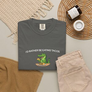 Peut inclure: Un t-shirt gris Comfort Colors avec le texte "I'D RATHER BE EATING TACOS" et un alligator de dessin animé mangeant des tacos. Le t-shirt est associé à un pantalon beige et à d'autres accessoires.
