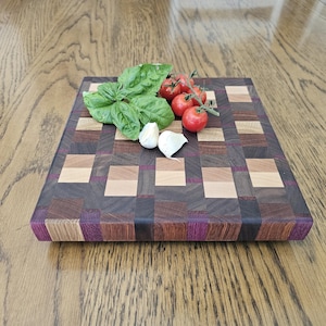 Può includere: Un tagliere artigianale in legno con un motivo a blocchi di diverse tonalità, con accenti viola. Basilico fresco, pomodori e spicchi d'aglio disposti sopra, suggerendo un uso culinario.