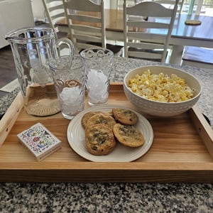 May include: A wooden serving tray holds a clear glass pitcher, two glasses with ice, a bowl of popcorn, a plate of cookies, and a deck of cards. The tray has a light wood grain and a rectangular shape.