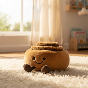 Puede incluir: Un juguete de peluche con forma de rollo de canela con una cara sonriente y pequeños brazos y pies marrones. El juguete suave de color canela está sobre una alfombra blanca en una habitación bien iluminada.