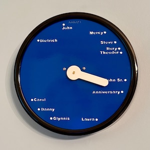 May include: A round, blue clock with a black frame. The clock face has white text with names like John, Carol, and Danny. A white, paddle-shaped hand points to the names. The word "Anniversary" is also visible.