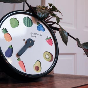 Op de afbeelding: Een ronde klok met een zwart frame en een witte wijzerplaat met kleurrijke illustraties van fruit en groenten. De wijzers zijn zwart en de tekst "grow baby, grow" staat op de wijzerplaat. Er staat een plant naast de klok.
