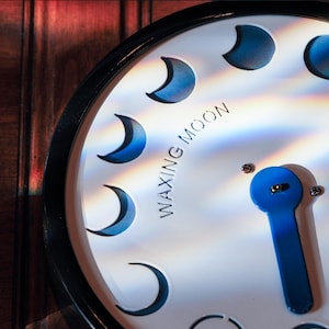 May include: A white clock face with a black border and a blue hand. The clock features crescent moon cutouts and the words "Waxing Moon". The clock is mounted on a wooden surface.