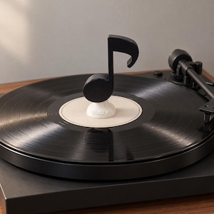 Pode incluir: Um estabilizador de discos em forma de nota musical preta com uma base branca contendo o texto SpinStyle. O estabilizador está posicionado no centro de um disco de vinil preto num gira-discos, que inclui um braço preto e uma base de madeira.