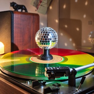 Puede incluir: Un estabilizador de discos en forma de bola de discoteca plateada con el texto SpinStyle impreso en su base negra. El estabilizador se sitúa en el centro de un disco de vinilo translúcido con segmentos rojos, amarillos y verdes en un tocadiscos.