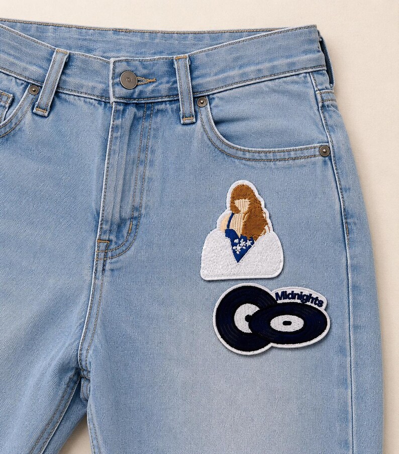 K&ouml;nnte beinhalten: Hellblaue Jeans aus Denim mit zwei gestickten Aufn&auml;hern auf der Vorderseite. Ein Aufn&auml;her zeigt eine Person mit langem braunen Haar in einem blauen Oberteil, der andere zeigt zwei schwarze Schallplatten mit dem in Blau gestickten Text Midnights.
