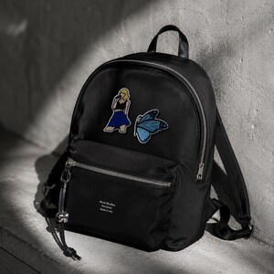 May include: A black backpack featuring a silver zip, a top handle, and adjustable shoulder straps. The front displays an embroidered patch of a person with blonde hair and a blue skirt, a blue butterfly patch, and the text Acne Studios Stockholm Made in Italy.