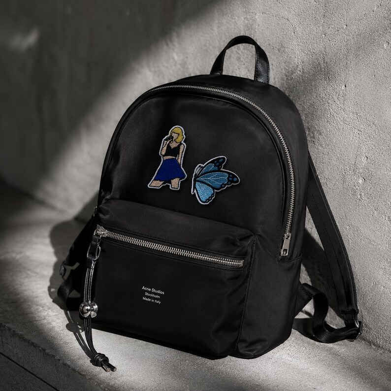 May include: A black backpack featuring a silver zip, a top handle, and adjustable shoulder straps. The front displays an embroidered patch of a person with blonde hair and a blue skirt, a blue butterfly patch, and the text Acne Studios Stockholm Made in Italy.