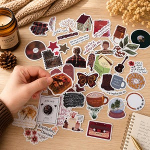 Pode incluir: Uma cole&ccedil;&atilde;o de autocolantes decorativos com v&aacute;rios desenhos, incluindo uma casa, uma borboleta, uma guitarra e texto. Os autocolantes apresentam uma mistura de cores, incluindo vermelho, castanho e branco. Alguns autocolantes t&ecirc;m texto como "evermore" e "gold rush".