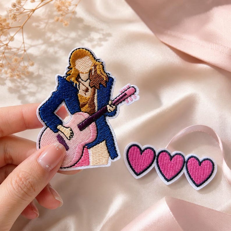 May include: An embroidered patch featuring a figure with blonde hair wearing a blue blazer and playing a pink acoustic guitar, alongside a separate embroidered patch consisting of three connected pink hearts with black outlines and white borders.