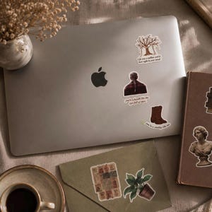 Pode incluir: Um laptop prateado adornado com v&aacute;rios adesivos, incluindo uma &aacute;rvore, uma pessoa e uma bota de cowboy. Os adesivos apresentam cita&ccedil;&otilde;es. Uma x&iacute;cara de caf&eacute;, um livro e um envelope com um adesivo de planta tamb&eacute;m s&atilde;o vis&iacute;veis.