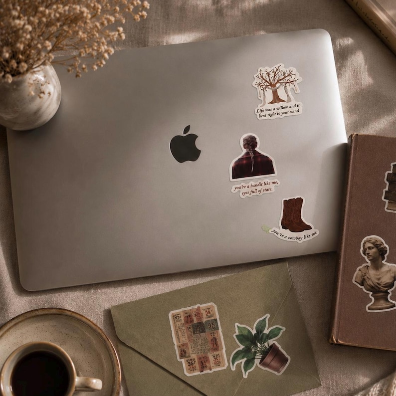 Pode incluir: Um laptop prateado adornado com v&aacute;rios adesivos, incluindo uma &aacute;rvore, uma pessoa e uma bota de cowboy. Os adesivos apresentam cita&ccedil;&otilde;es. Uma x&iacute;cara de caf&eacute;, um livro e um envelope com um adesivo de planta tamb&eacute;m s&atilde;o vis&iacute;veis.