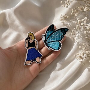 May include: Two embroidered patches: one depicts a blonde figure in a black top and blue skirt holding a microphone, and the other is a blue and black butterfly with intricate wing patterns, both featuring white borders and detailed stitching.