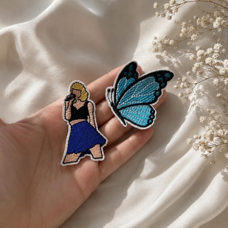 May include: Two embroidered patches: one depicts a blonde figure in a black top and blue skirt holding a microphone, and the other is a blue and black butterfly with intricate wing patterns, both featuring white borders and detailed stitching.