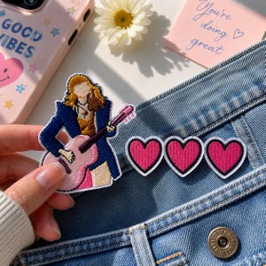 May include: An embroidered patch featuring a person with long blonde hair wearing a dark blue jacket and tan pants while playing a light pink acoustic guitar, accompanied by three separate heart-shaped patches with pink centers and white borders.