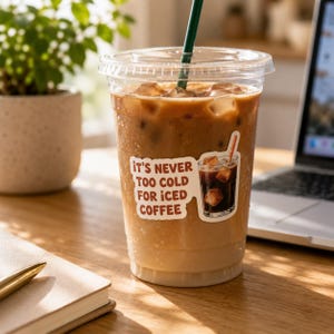 Peut inclure: Un gobelet en plastique transparent rempli de café glacé, de glaçons et d'une paille verte. Une étiquette sur le gobelet indique "It's never too cold for iced coffee". Le gobelet est posé sur une surface en bois.
