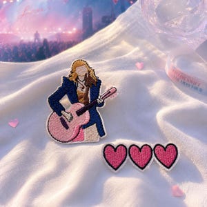 May include: An embroidered patch featuring a figure with blonde hair wearing a dark blue jacket and playing a pink acoustic guitar, accompanied by a separate embroidered patch consisting of three connected pink hearts with black outlines.