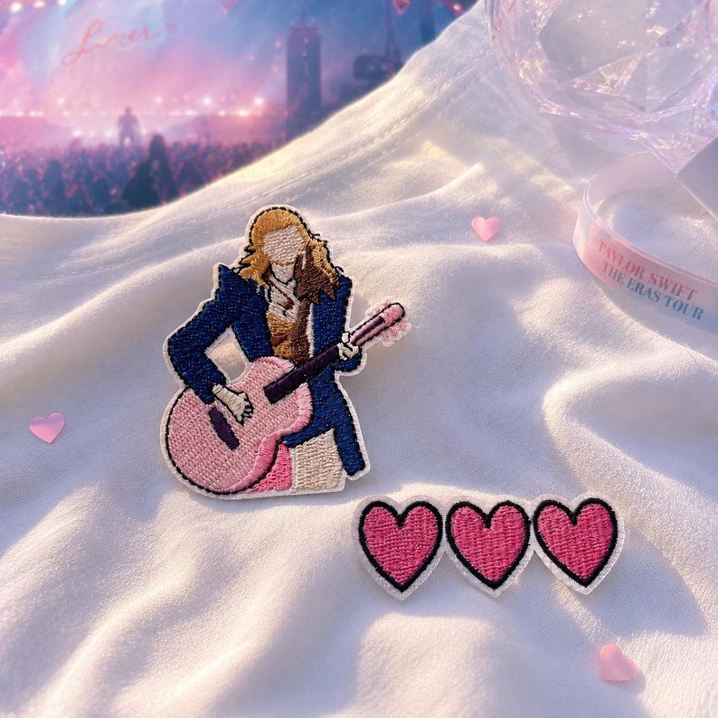 May include: An embroidered patch featuring a figure with blonde hair wearing a dark blue jacket and playing a pink acoustic guitar, accompanied by a separate embroidered patch consisting of three connected pink hearts with black outlines.