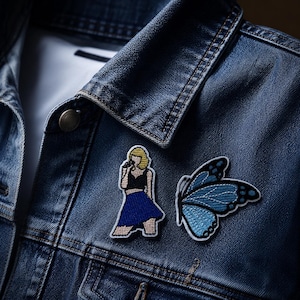 May include: A dark blue denim jacket featuring two embroidered patches. One patch depicts a figure with blonde hair wearing a black top and a blue skirt while holding a microphone. The second patch is a blue butterfly with black wing details.