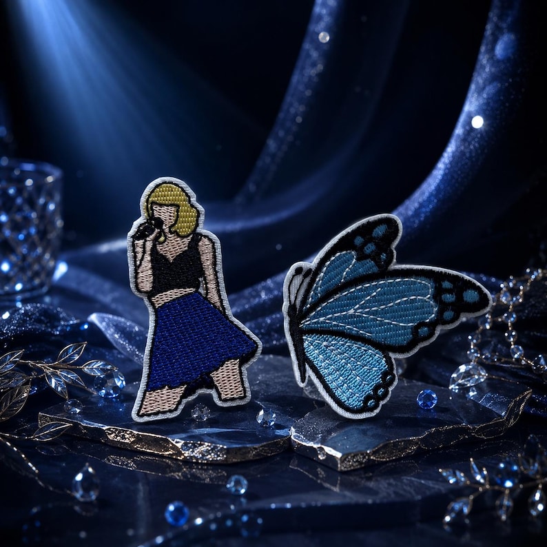 May include: Two embroidered patches featuring a figure with blonde hair wearing a black top and blue skirt holding a microphone, and a blue butterfly with black wing edges and white details. The patches are displayed on dark, textured surfaces.