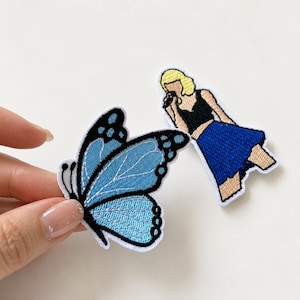 May include: Two embroidered patches featuring a light blue butterfly with black outlines and a stylised figure with blonde hair wearing a black top and a blue skirt while holding a microphone. The patches have white borders and detailed stitching.