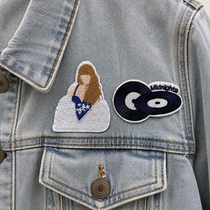 K&ouml;nnte beinhalten: Eine hellblaue Jeansjacke mit zwei gestickten Aufn&auml;hern. Ein Aufn&auml;her zeigt eine Person mit langem braunen Haar in einem blauen Oberteil mit wei&szlig;en Sternen, der andere zeigt zwei schwarze Schallplatten mit dem wei&szlig;en Schriftzug Midnights.