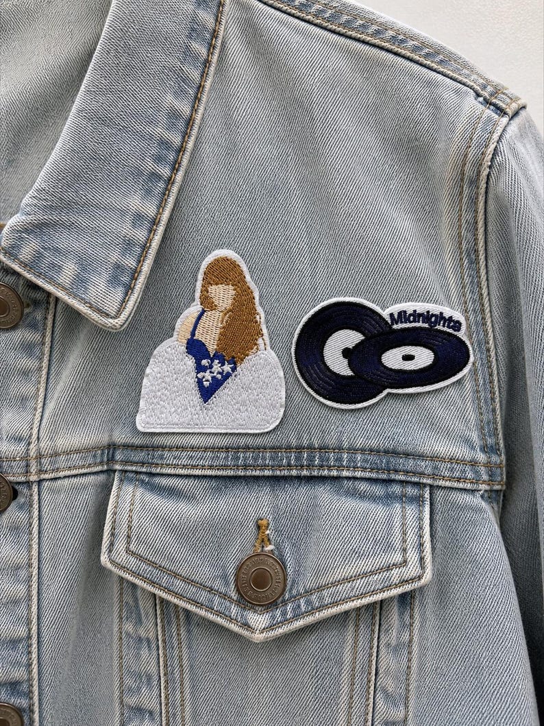 K&ouml;nnte beinhalten: Eine hellblaue Jeansjacke mit zwei gestickten Aufn&auml;hern. Ein Aufn&auml;her zeigt eine Person mit langem braunen Haar in einem blauen Oberteil mit wei&szlig;en Sternen, der andere zeigt zwei schwarze Schallplatten mit dem wei&szlig;en Schriftzug Midnights.